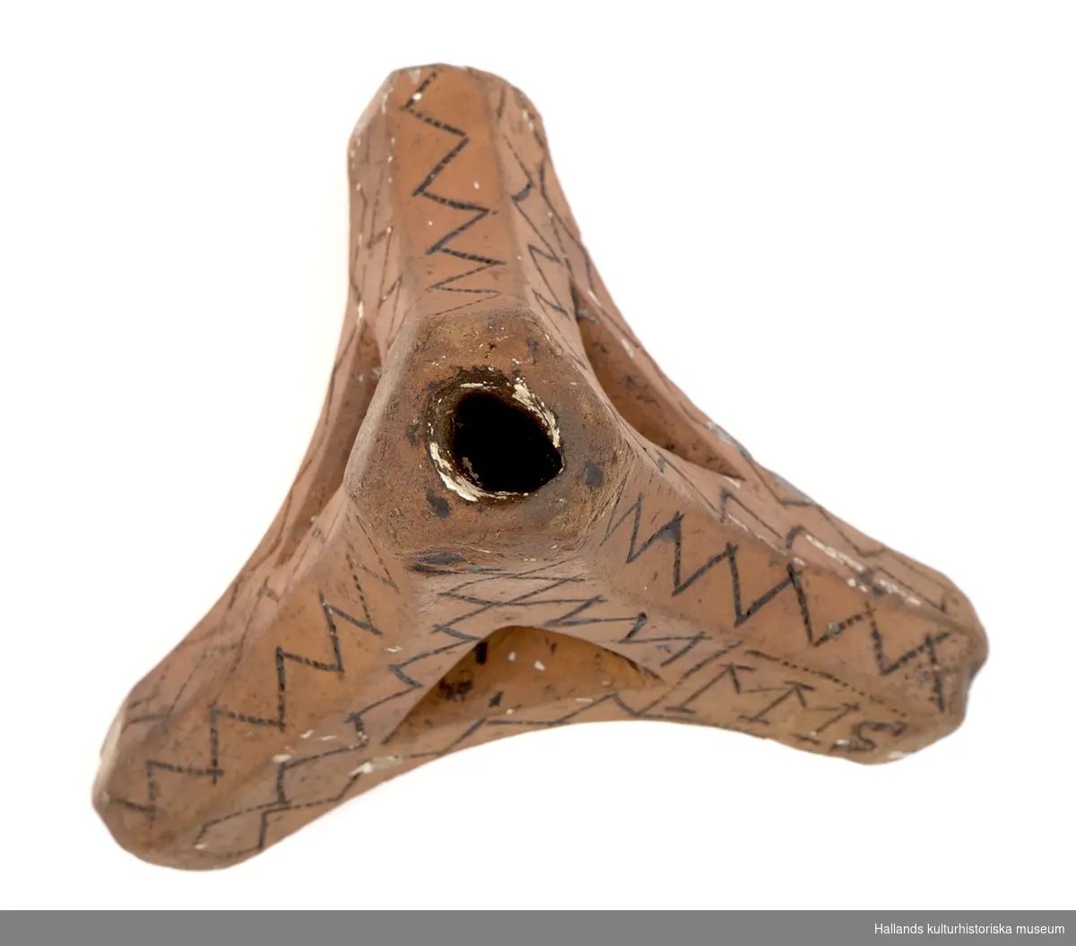 Ljusstake, en s.k, "Fotangelstake", av bränt lergods. Triangulär form, består av fyra pipor. Rikligt dekorerad med präglade sicksackmönster som förstärkts med sotning. I mönstret en signering: "MMS".