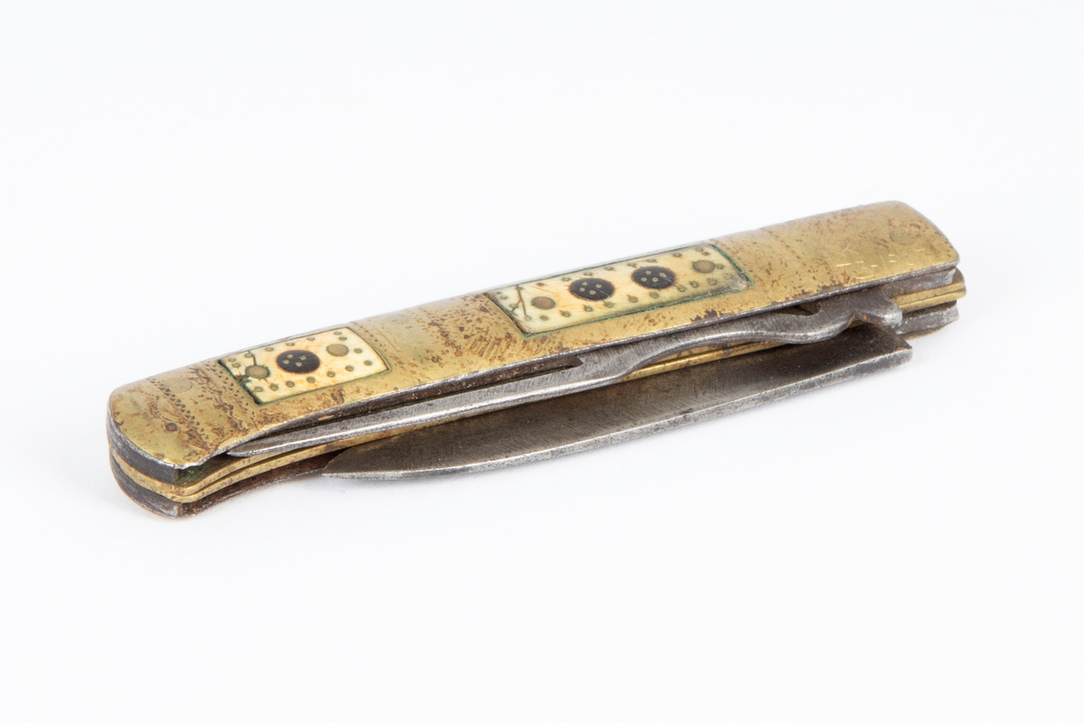 Foldekniv med kniv og gaffel. Kniven er spiss og har nærmest lansettform. Gaffelen rett og flat med 3 tenner og gjennombrutt hjerteform nær roten. Skaftet består av to sammenføyde messingplater som på hver side har innlagt rektangulære felt av horn igjen innlagt med messing. Siselert dekor på messingskaftet med buer og prikker.