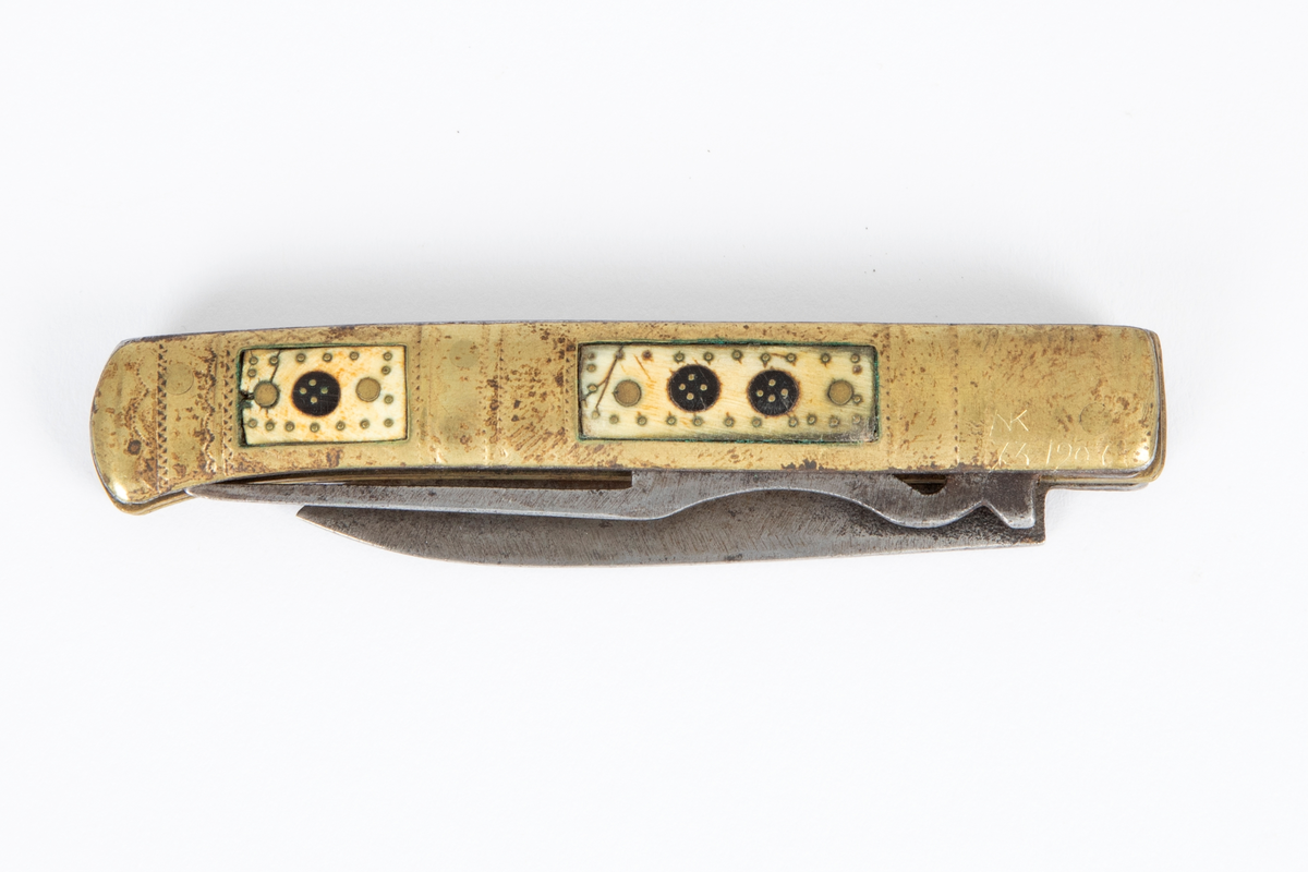 Foldekniv med kniv og gaffel. Kniven er spiss og har nærmest lansettform. Gaffelen rett og flat med 3 tenner og gjennombrutt hjerteform nær roten. Skaftet består av to sammenføyde messingplater som på hver side har innlagt rektangulære felt av horn igjen innlagt med messing. Siselert dekor på messingskaftet med buer og prikker.
