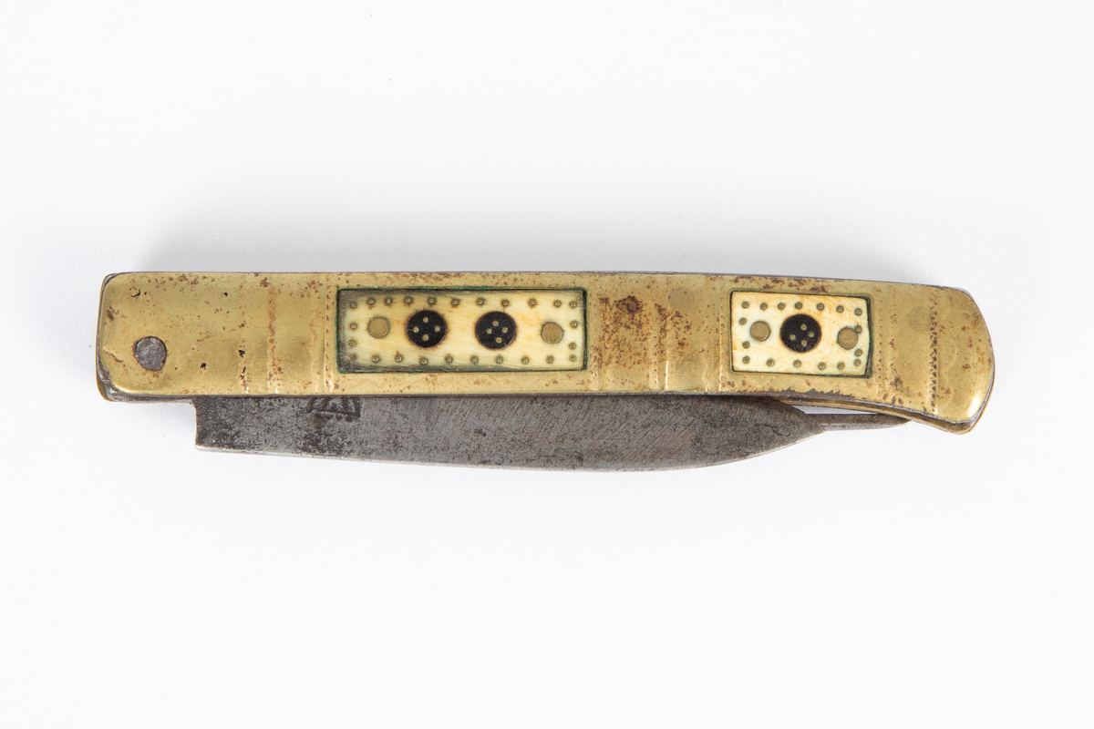 Foldekniv med kniv og gaffel. Kniven er spiss og har nærmest lansettform. Gaffelen rett og flat med 3 tenner og gjennombrutt hjerteform nær roten. Skaftet består av to sammenføyde messingplater som på hver side har innlagt rektangulære felt av horn igjen innlagt med messing. Siselert dekor på messingskaftet med buer og prikker.