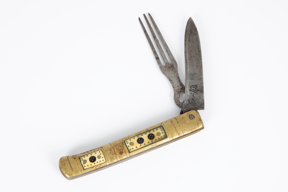 Foldekniv med kniv og gaffel. Kniven er spiss og har nærmest lansettform. Gaffelen rett og flat med 3 tenner og gjennombrutt hjerteform nær roten. Skaftet består av to sammenføyde messingplater som på hver side har innlagt rektangulære felt av horn igjen innlagt med messing. Siselert dekor på messingskaftet med buer og prikker.