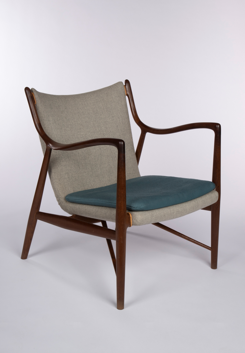 Stol i åpen, lett konstruksjon med vertikalstilte forben, og sterkt skråstilte bakben. Mykt svungne armlener festet med jevn overgang til for- og bakben, og som herfra er svunget opp som akterstaver. Disse er forbundet med et ryggbrett som er skjult av stopningen. Overgangene på sidene er kantet med lyst lær. Mellom de to sidesargene, som også fungerer som sidesprosser, et midtstykke som støtte for setet. Dreiede sprosser løpter fra nederst på forbenene på skrå opp til midtstykket. Mellom forbenene er forsargen skjult av stopningen og kantet med lyst lær på hver side. Sammenhengende sete- og ryggstopning, trukket med grått ullstoff med løs pute i blågrønt ullstoff.