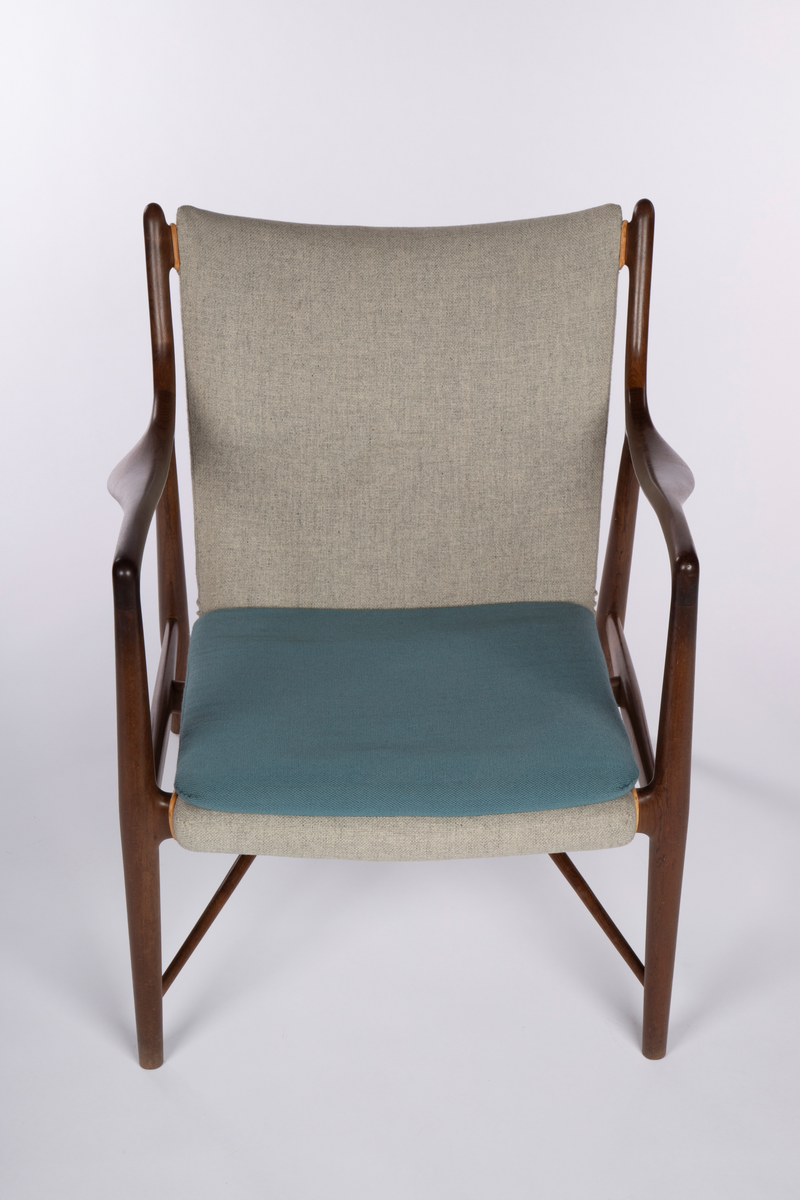 Stol i åpen, lett konstruksjon med vertikalstilte forben, og sterkt skråstilte bakben. Mykt svungne armlener festet med jevn overgang til for- og bakben, og som herfra er svunget opp som akterstaver. Disse er forbundet med et ryggbrett som er skjult av stopningen. Overgangene på sidene er kantet med lyst lær. Mellom de to sidesargene, som også fungerer som sidesprosser, et midtstykke som støtte for setet. Dreiede sprosser løpter fra nederst på forbenene på skrå opp til midtstykket. Mellom forbenene er forsargen skjult av stopningen og kantet med lyst lær på hver side. Sammenhengende sete- og ryggstopning, trukket med grått ullstoff med løs pute i blågrønt ullstoff.