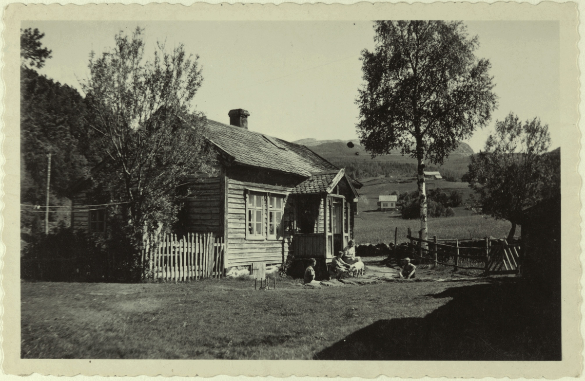 Eit eldre trehus i eit jordbrukslandskap. Det sit fire personar på og ved trappa til huset, og dei har ein gris med seg.