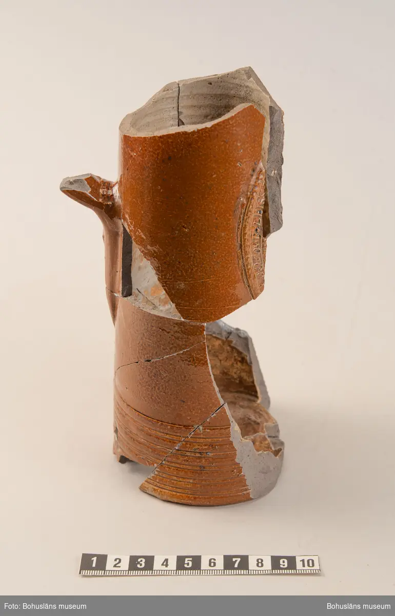 Krus/Kanna. Stort brunglaserat stengodskärl med dekor i form av delvis bevarad textmedaljong på framsida kanna. Tysk text. 

Landskap: Bohuslän