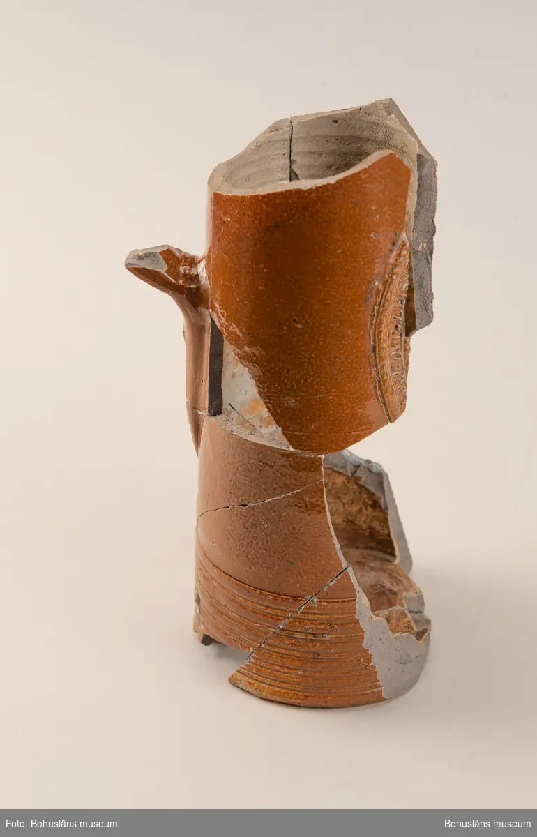 Krus/Kanna. Stort brunglaserat stengodskärl med dekor i form av delvis bevarad textmedaljong på framsida kanna. Tysk text. 

Landskap: Bohuslän