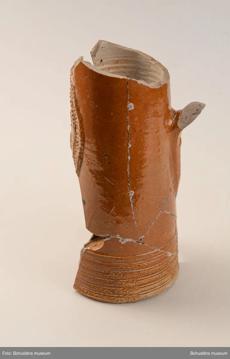 Krus/Kanna. Stort brunglaserat stengodskärl med dekor i form av delvis bevarad textmedaljong på framsida kanna. Tysk text. 

Landskap: Bohuslän