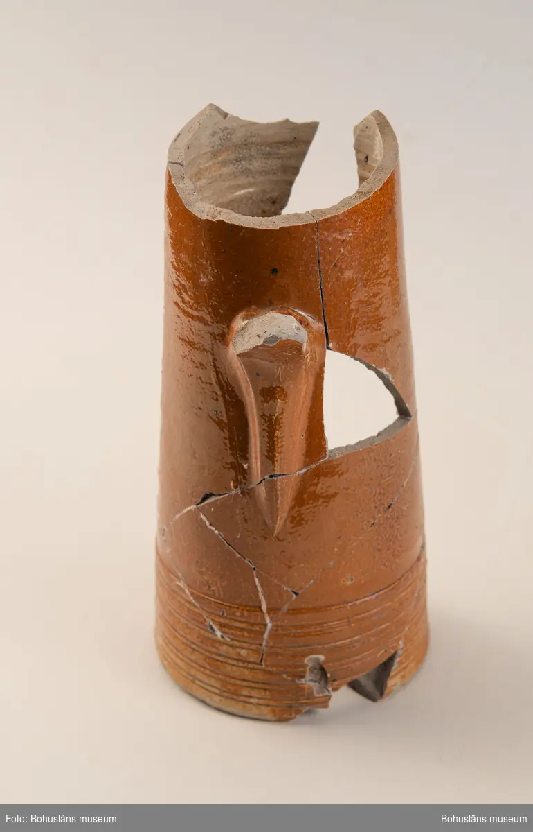 Krus/Kanna. Stort brunglaserat stengodskärl med dekor i form av delvis bevarad textmedaljong på framsida kanna. Tysk text. 

Landskap: Bohuslän