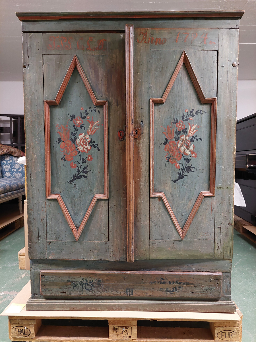 Skåp i allmogestil med dubbeldörrar och en låda.

Skåpet är målat i en blågrön färg och dekorerad på dörrspeglarna och lådan. Dörrspeglarna har profilerade rödmålade lister. I speglarna är det målade blommor och blad i vita, röda och blå färger. Lådan är dekorerad med blad i blå färg. 

Överst på dörrarna finns ett årtal, 1794, samt initialer målade i rött.

Inuti skåpet fins tre hyllpan, det översta med utskurna hack för skedar, en så kallad skedhylla. En stor låda finns under dörrarna.

Dörrarna är försedda med smidda gångjärn och två hjärtformade nyckelskyltar, det vänstra är ett blindhål.
