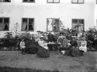 Makarna August Fredholm och Helga Cavallin med barn samlade för fotografering. Platsen är makarnas gods Karlshov i Älvestad och tidpunkten sannolikt i samband med Fredholms dotter Ingrids bröllop i september 1896. Vi ser henne sittande på bänken till höger med fadern vid sin sida. De mindre barnen är ättlingar till den omgifte fadern och nämnda Helga Cavallin, som vi ser sitta i förgrunden. I knäet har hon makarnas sistfödde, Nils. Parets övriga barn ses från vänster i Einar, Signe (vid fadern), Jane (vid Ingrid) och Hildur längst till höger på bänken. De bägge stående personerna och kvinnan i mörkt på bänken har inte identifierats.