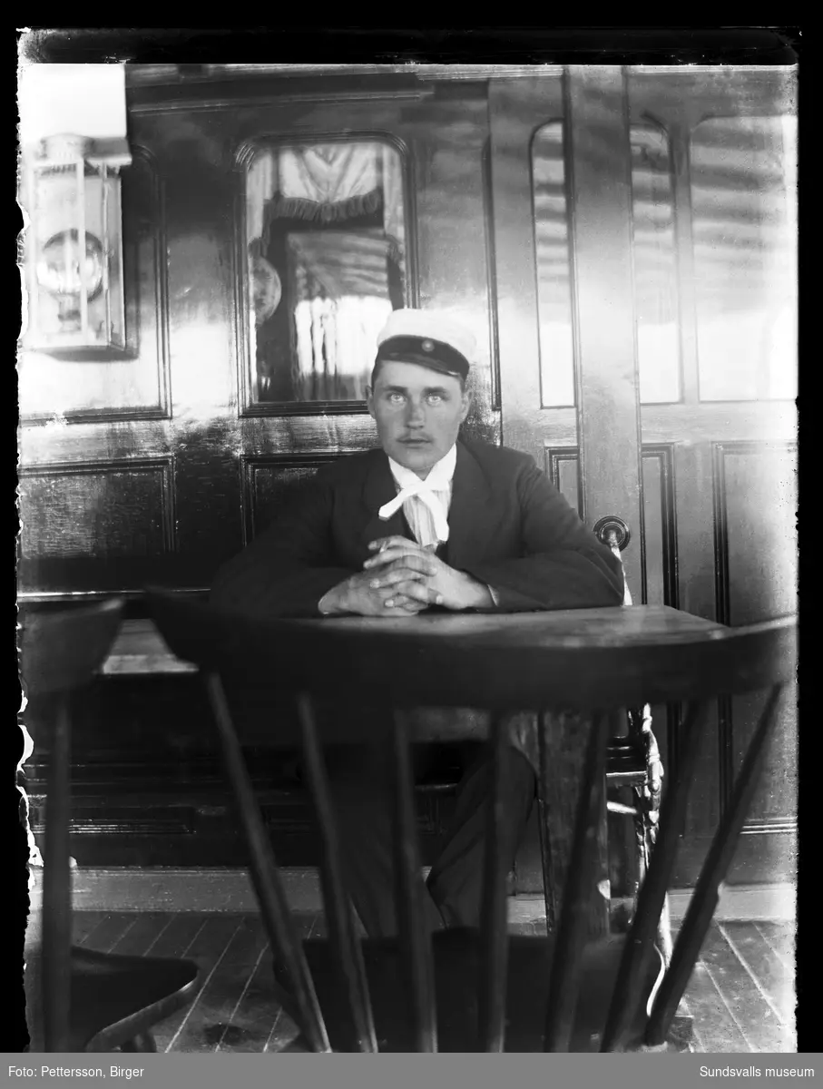 Porträttbild med en man som sitter vid ett bord i ett rum med mörk träpanel och fönsterdörrar.