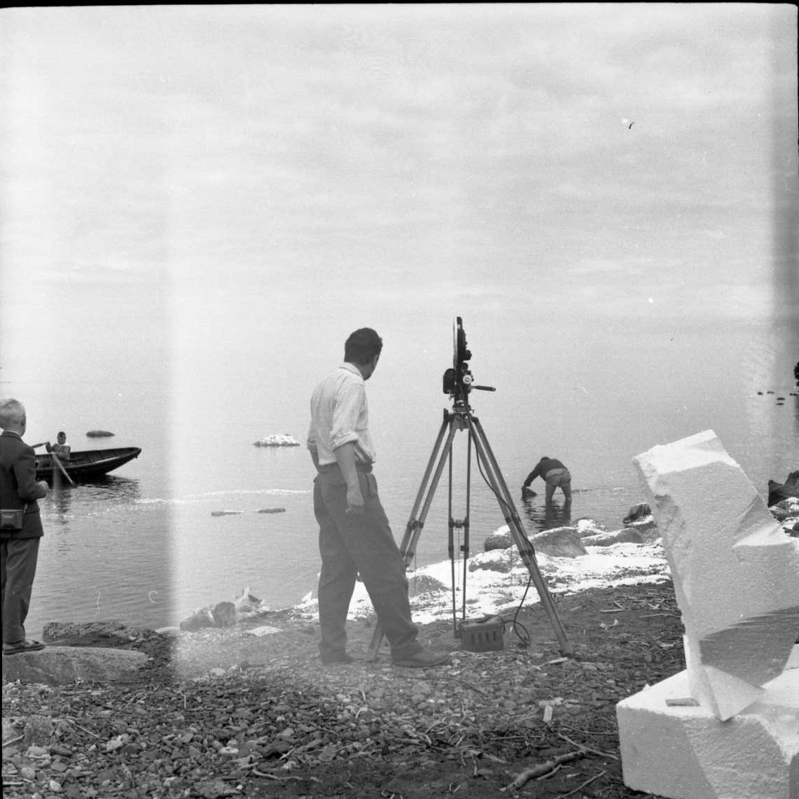 Ett filmteam vid Vätterstranden, norr om hamne i Gränna, lite publik i bakgrunden. Konstgjord snö på stranden. En roddbåt är på väg mot stranden.