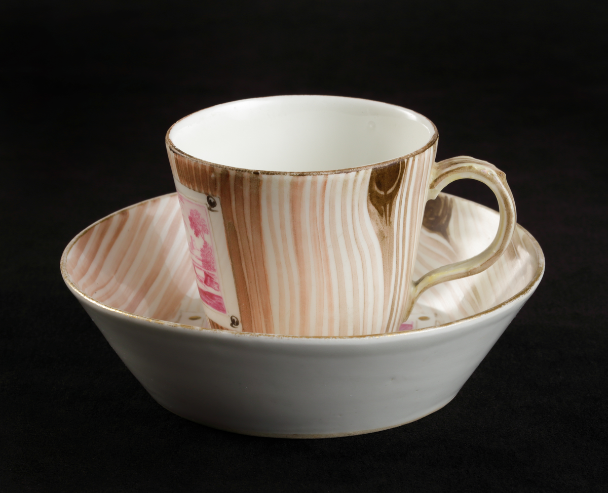 Kopp med fat av porslin. Rak bägarform med C-formad grepe. Fat med raka kanter. 
Dekorerad i träådring, med simulerade pappersblad (trompe l´oeil) och landskap i rött efter
kopparstick. Kantränder i brunt och guld.

Inskrivet i huvudkatalogen 1967.