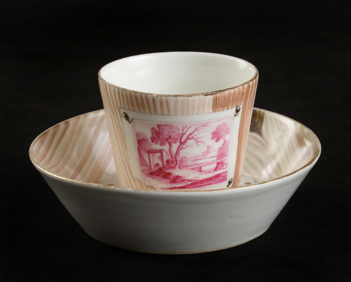 Kopp med fat av porslin. Rak bägarform med C-formad grepe. Fat med raka kanter. 
Dekorerad i träådring, med simulerade pappersblad (trompe l´oeil) och landskap i rött efter
kopparstick. Kantränder i brunt och guld.

Inskrivet i huvudkatalogen 1967.