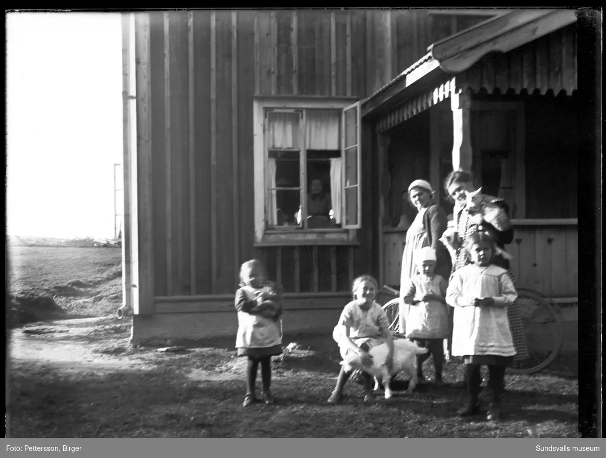 Framför ett lantligt beläget bostadshus står två kvinnor och fyra barn, i fönstrer bakom syns ytterligare en kvinna. Med på bilden är även tre katter och en get.