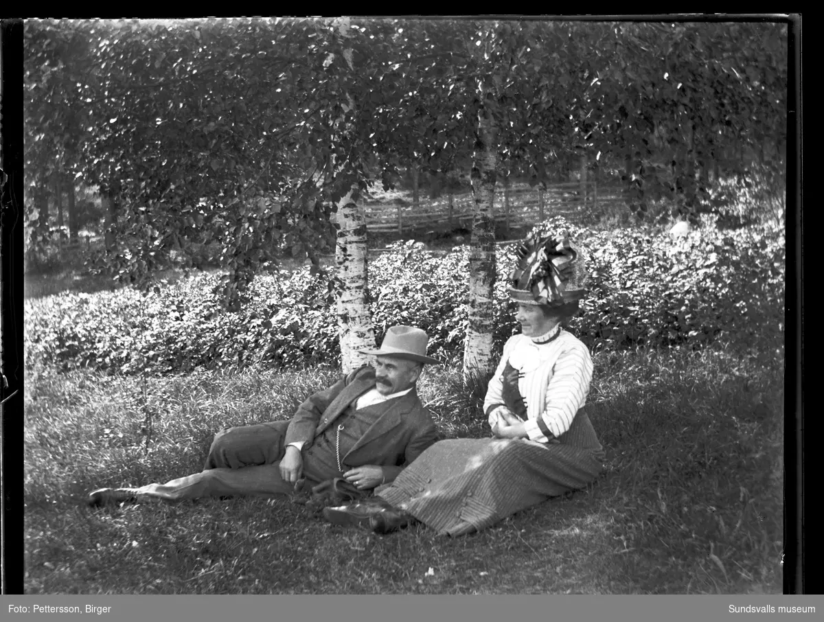 En man och en kvinna i gräset invid två björkar i en trädgård eller park. Kvinnan har en rikt dekorerad och hög hatt.