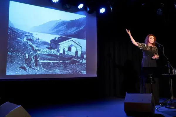 Forestillingsbilde. Sangerinnen til høyre i sort kjole viser til bildet på et lerret til venstre. Bildet er et gammelt landskapsfoto med to hus langs et vann med fjell i bakgrunn. Fo mennesker i turklær med sekk og staver i forgrunnen. To menn og to damer står utenfor huset. 