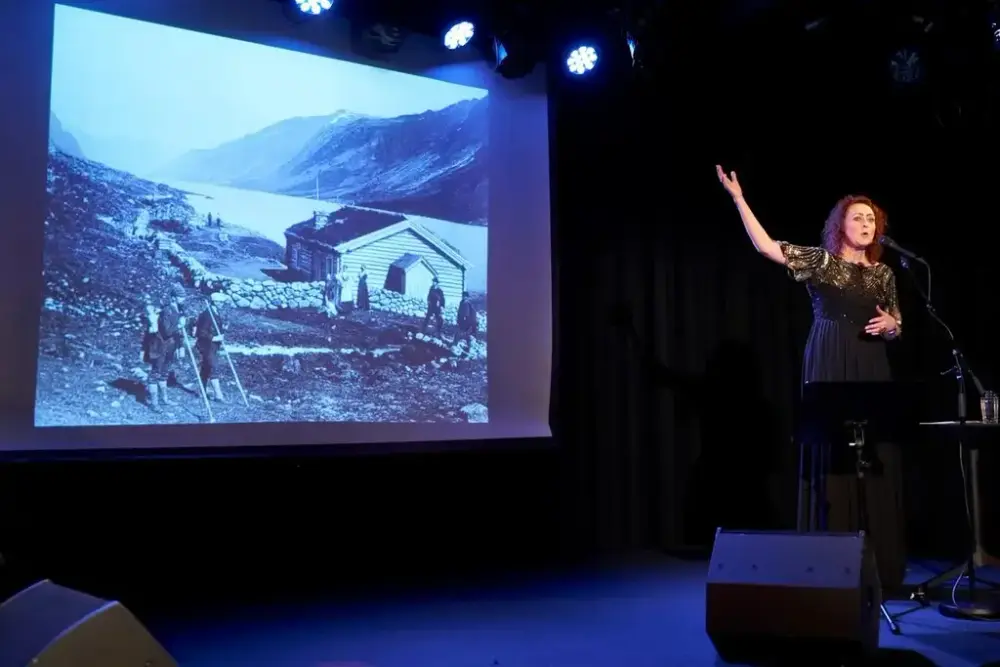 Forestillingsbilde. Sangerinnen til høyre i sort kjole viser til bildet på et lerret til venstre. Bildet er et gammelt landskapsfoto med to hus langs et vann med fjell i bakgrunn. Fo mennesker i turklær med sekk og staver i forgrunnen. To menn og to damer står utenfor huset. 