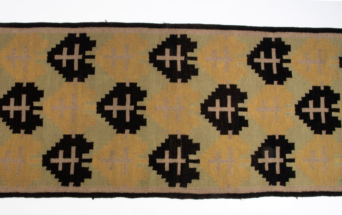 Rektangulær gulvløper i rutevevteknikk i tykt ullgarn. Innslag i svart, gult, grønt og beige på grov hamprenning. Motivet er et stilisert tre eller "hjerte" i svart og ett i gult; gjentatt regelmessig langs hele løperen.
