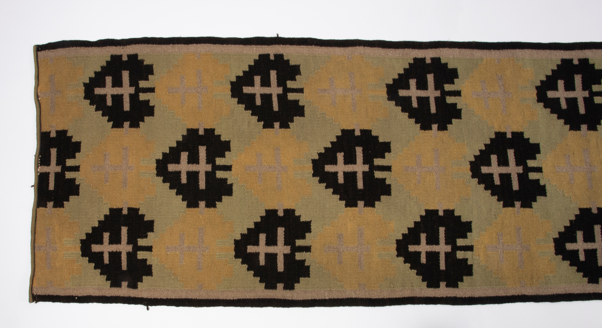 Rektangulær gulvløper i rutevevteknikk i tykt ullgarn. Innslag i svart, gult, grønt og beige på grov hamprenning. Motivet er et stilisert tre eller "hjerte" i svart og ett i gult; gjentatt regelmessig langs hele løperen.
