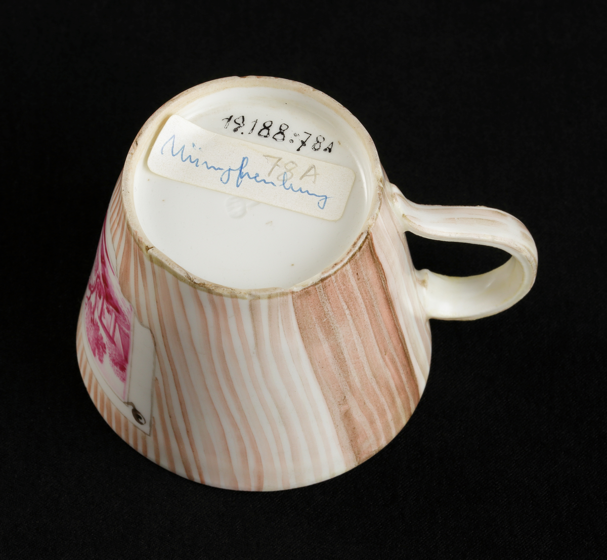 Kopp med fat av porslin. Rak bägarform med C-formad grepe. Fat med raka kanter. 
Dekorerad i träådring, med simulerade pappersblad (trompe l´oeil) och landskap i rött efter
kopparstick. Kantränder i brunt och guld.

Inskrivet i huvudkatalogen 1967.