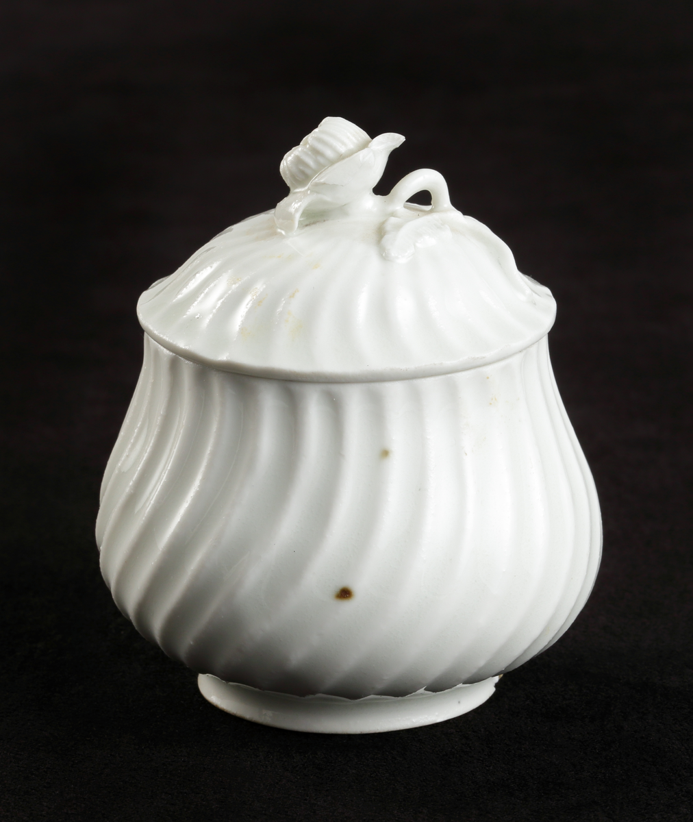 Krämkopp med lock, av porslin. Vitglaserad.
Något bukig modell, vridna räfflor å lock och knopp. På locket en skulpterad blomma som knopp.

Inskrivet i huvudbok 1967.