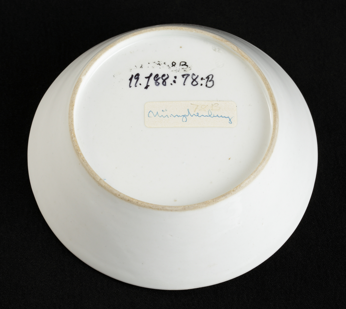 Kopp med fat av porslin. Rak bägarform med C-formad grepe. Fat med raka kanter. 
Dekorerad i träådring, med simulerade pappersblad (trompe l´oeil) och landskap i rött efter
kopparstick. Kantränder i brunt och guld.

Inskrivet i huvudkatalogen 1967.