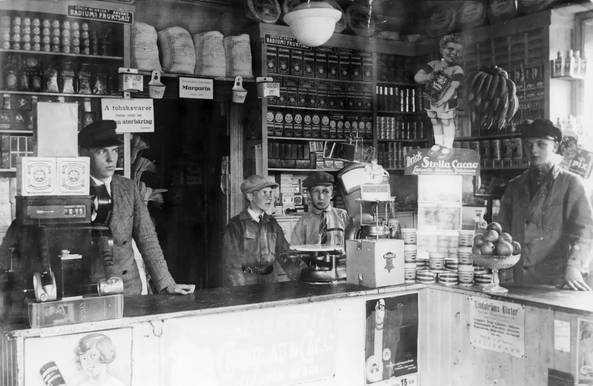 Interiör från Rönninge andelsförenings (Konsum) filial åren 1928-1929.

Affären låg i Engelsberg, Fiskaruddsvägen 2, byggt år 1905. Från vänster står affärsföreståndaren Yngve Carell, obekant, Åke Abrahamsson (halvbror till konstnären Hans Abrahamsson) och Harry Jonsson. Lägg märke till reklamskylten längst upp till vänster: "Söker ni hälsa? Använd RADIUMs FRUKTSALT". 

Yngve Carell övertog affären som från år 1935 och under 20-25 år innehades av Alex Malmberg. Förutom speceri- och charkuterivaror såldes även järnvaror, fönsterglas och byggnadsmaterial. När verksamheten så småningom upphörde och hyresgästerna flyttat förföll huset, för att på 1990-talet rustas upp och renoveras till hyreslägenheter.

Uppgiftslämnare: Anders Ahlberg 1997.