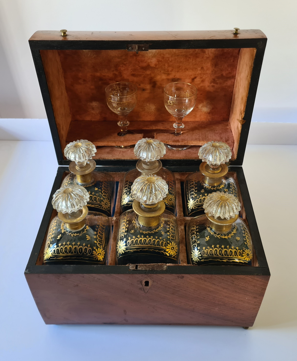 Reiseskrin med seks karafler og to glass. Skrinet er utformet i lys mahogny. Plass innvendig til seks karafler med propp, og to små stettglass. Skrinet er inndelt i seks seksjoner og trukket med mørk blå fløyel nede i rommene og (blass) rød fløyel oppe ved kantene og under lokket. I lokket er det også et spor til å feste to glass. Disse er det ikke plass til når skrinet lukkes. Hengslet lokk. Nøkkelhull foran. Nøkkel mangler.