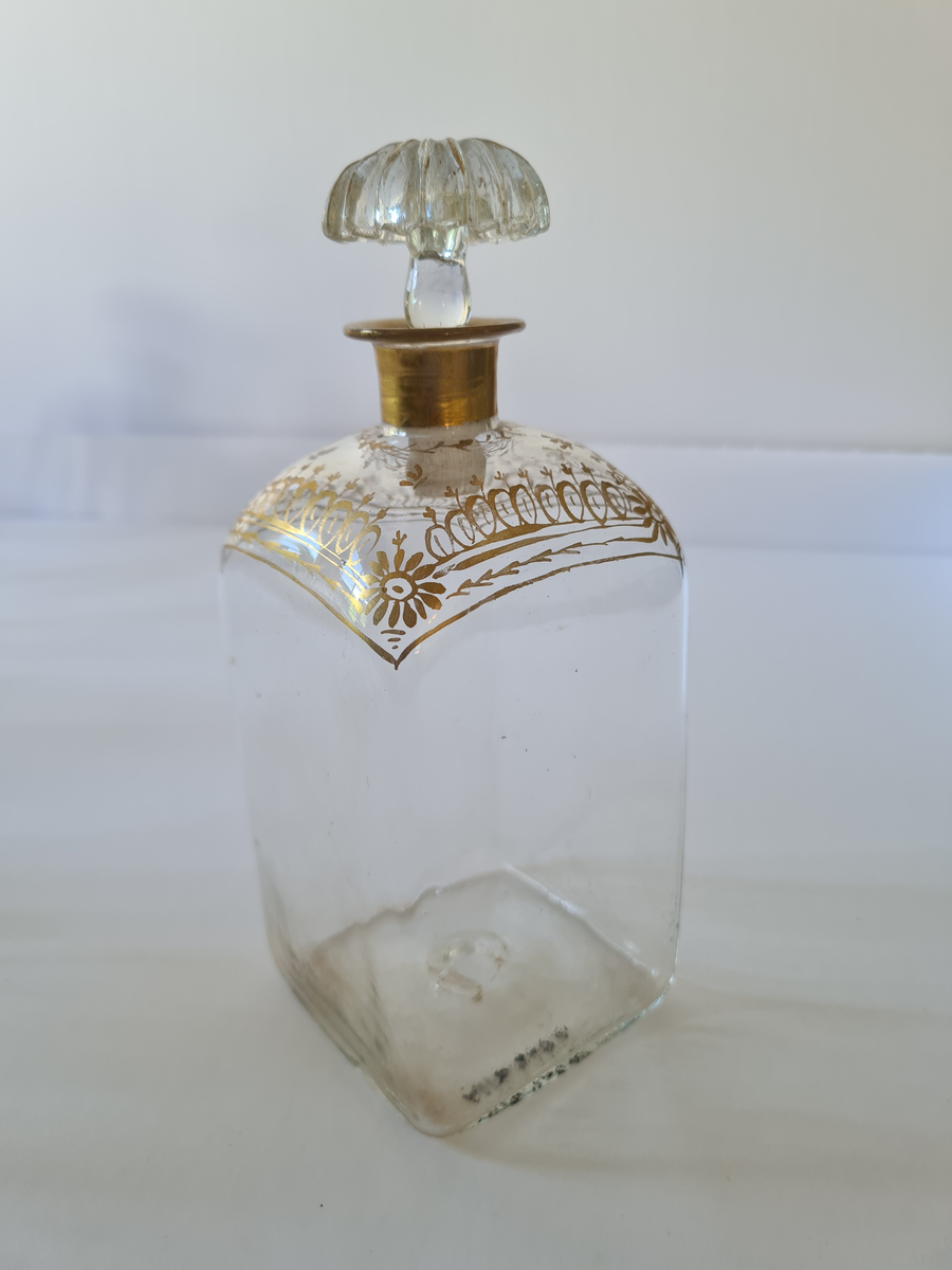 Karaffel av glass med tilhørende propp. Forgyllet dekor rundt halsen. Firkantet form, noe avrundet ved halsen.