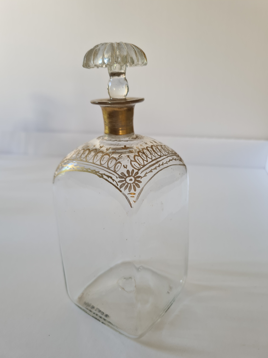 Karaffel av glass med tilhørende propp. Forgyllet dekor rundt halsen. Firkantet form, noe avrundet ved halsen.