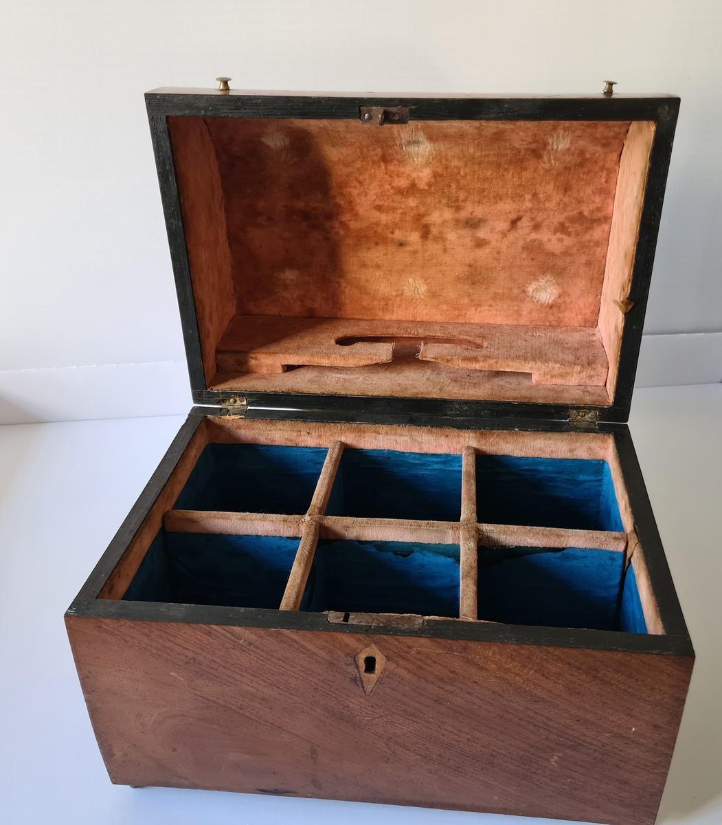 Reiseskrin med seks karafler og to glass. Skrinet er utformet i lys mahogny. Plass innvendig til seks karafler med propp, og to små stettglass. Skrinet er inndelt i seks seksjoner og trukket med mørk blå fløyel nede i rommene og (blass) rød fløyel oppe ved kantene og under lokket. I lokket er det også et spor til å feste to glass. Disse er det ikke plass til når skrinet lukkes. Hengslet lokk. Nøkkelhull foran. Nøkkel mangler.