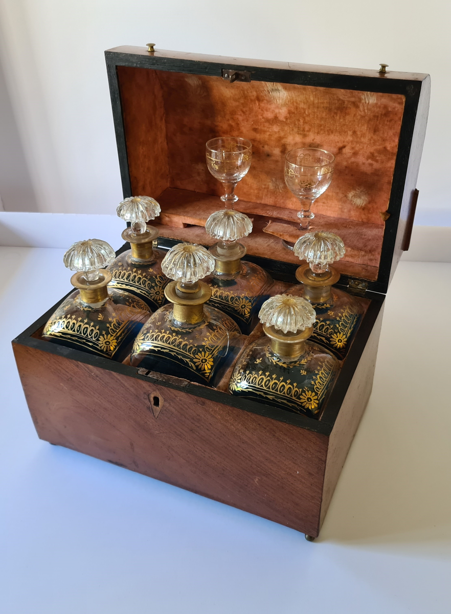 Reiseskrin med seks karafler og to glass. Skrinet er utformet i lys mahogny. Plass innvendig til seks karafler med propp, og to små stettglass. Skrinet er inndelt i seks seksjoner og trukket med mørk blå fløyel nede i rommene og (blass) rød fløyel oppe ved kantene og under lokket. I lokket er det også et spor til å feste to glass. Disse er det ikke plass til når skrinet lukkes. Hengslet lokk. Nøkkelhull foran. Nøkkel mangler.
