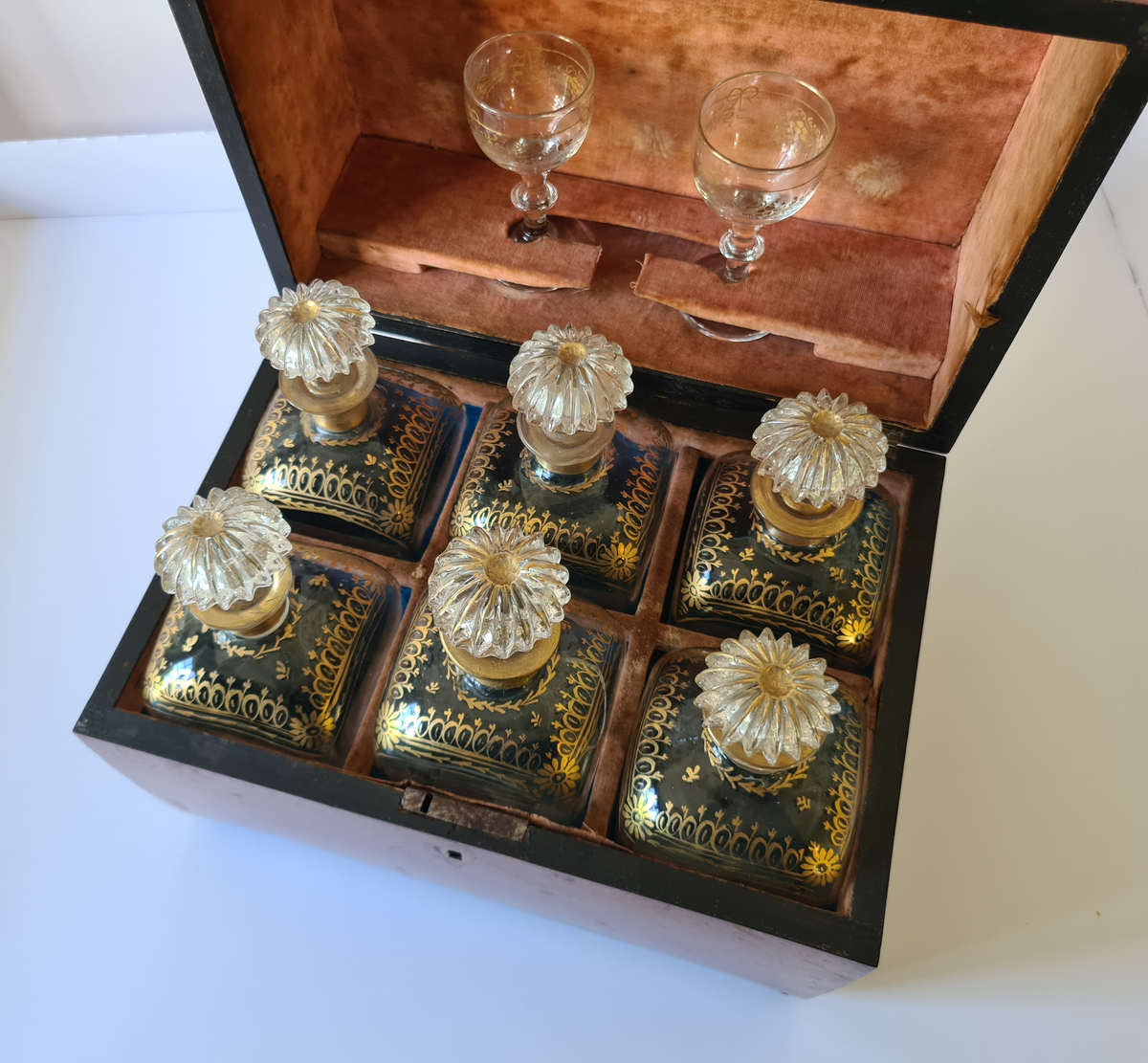 Reiseskrin med seks karafler og to glass. Skrinet er utformet i lys mahogny. Plass innvendig til seks karafler med propp, og to små stettglass. Skrinet er inndelt i seks seksjoner og trukket med mørk blå fløyel nede i rommene og (blass) rød fløyel oppe ved kantene og under lokket. I lokket er det også et spor til å feste to glass. Disse er det ikke plass til når skrinet lukkes. Hengslet lokk. Nøkkelhull foran. Nøkkel mangler.