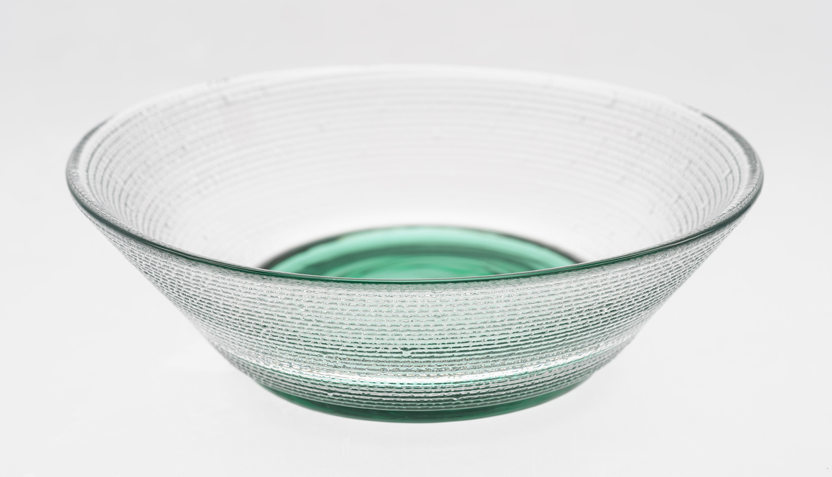 Konisk skål i gjennomskinnelig polykromt glass. Korpus er utført i klart glass, som glir over i en grønnfarget bunn. Yttersiden er dekket med horisontale riller, mens innsiden er glatt.