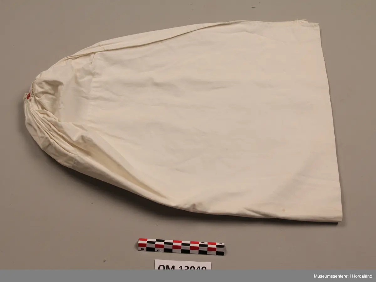 Kvithue av bomullslerret. Rynking i eine enden, med brodert rosett. Dobbelt stoff i enden mot hovudet. Handsauma.