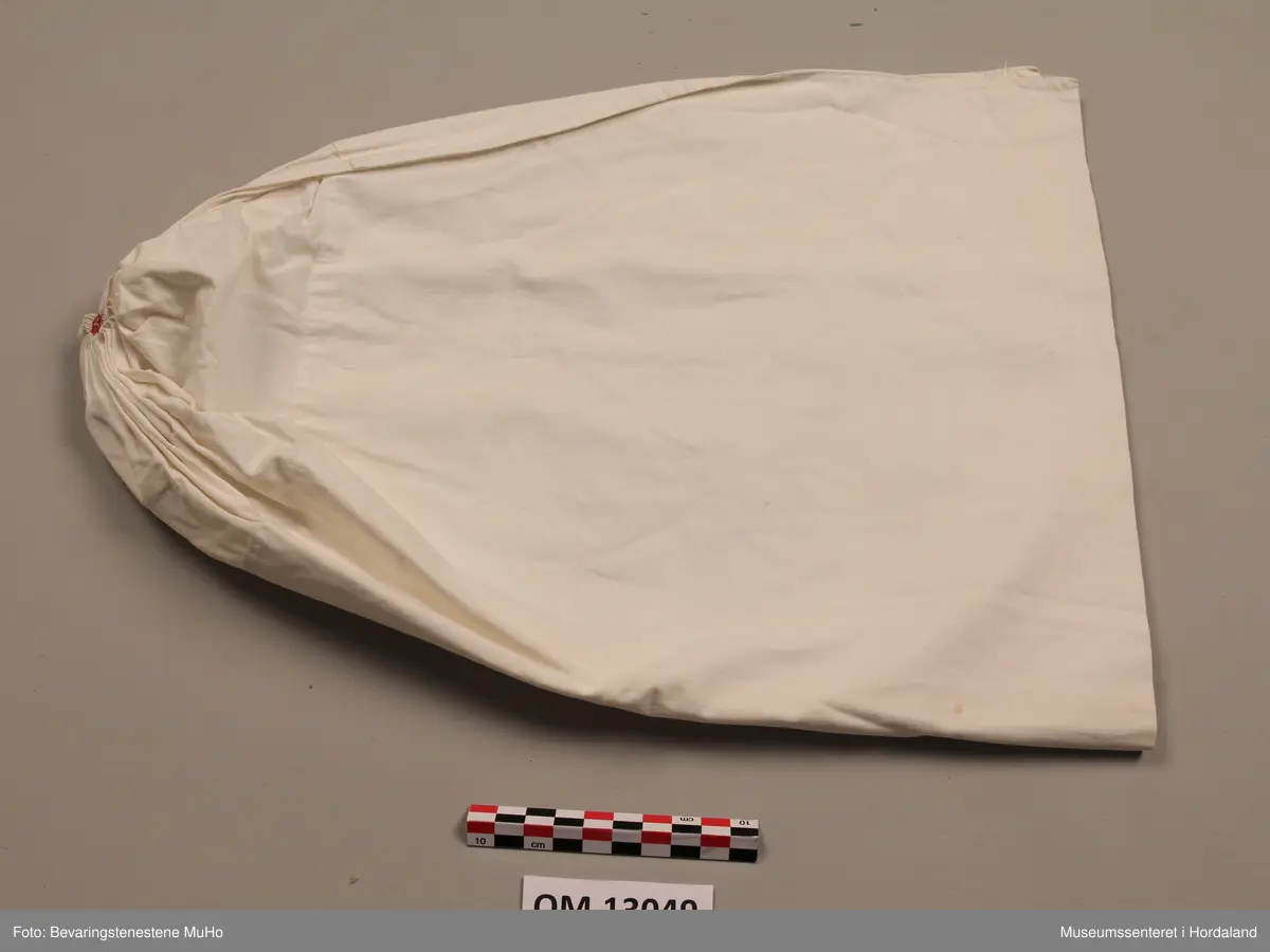 Kvithue av bomullslerret. Rynking i eine enden, med brodert rosett. Dobbelt stoff i enden mot hovudet. Handsauma.