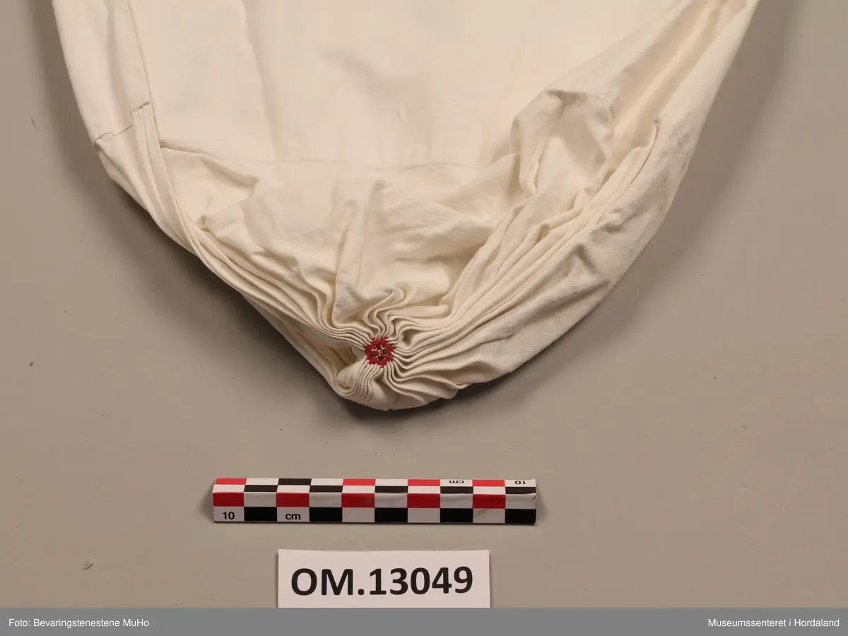 Kvithue av bomullslerret. Rynking i eine enden, med brodert rosett. Dobbelt stoff i enden mot hovudet. Handsauma.