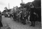 Personer utklädda som historiska personer, till exempel Gustav Vasa, i täten framför ett led med barn som bär blomsterbågar. De står på Brahegatan i Gränna.