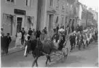 Kortege på Brahegatan i Gränna med häst och vagn i täten följt av män i folkdräkt. Det är hembygdsfest.