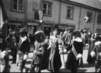 Kortege på Brahegatan i Gränna. Män och kvinnor i historiska dräkter. Fasadskyltar för 