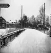 Längst en snötäckt väg sträcker sig ett staket. Bortanför staketet står avlövade träd och hus med snötäckta tak. Uppe i vänstra hörnet på bilden syns en liten del av en skylt där det står 