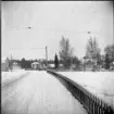 Längst en snötäckt väg sträcker sig ett staket. Bortanför staketet står hus med snötäckta tak och avlövade träd.