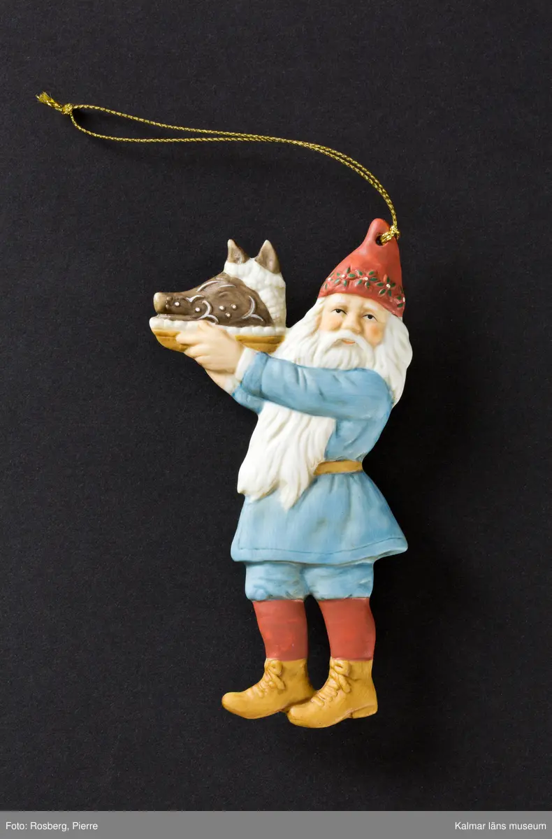 En tomte klädd i röd luva, blå jacka, blå knäkorta byxor, röda strumpor och bruna skor. Han håller upp ett fat med ett griljerat grishuvud.