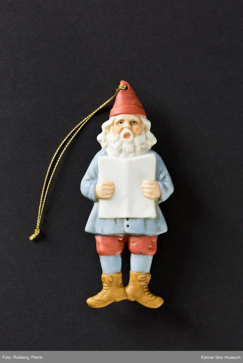 En tomte klädd i röd luva, blå jacka, röda knäkorta byxor, blå strumpor och bruna skor. Han håller i en bok eller nothäfte med båda händerna och sjunger.