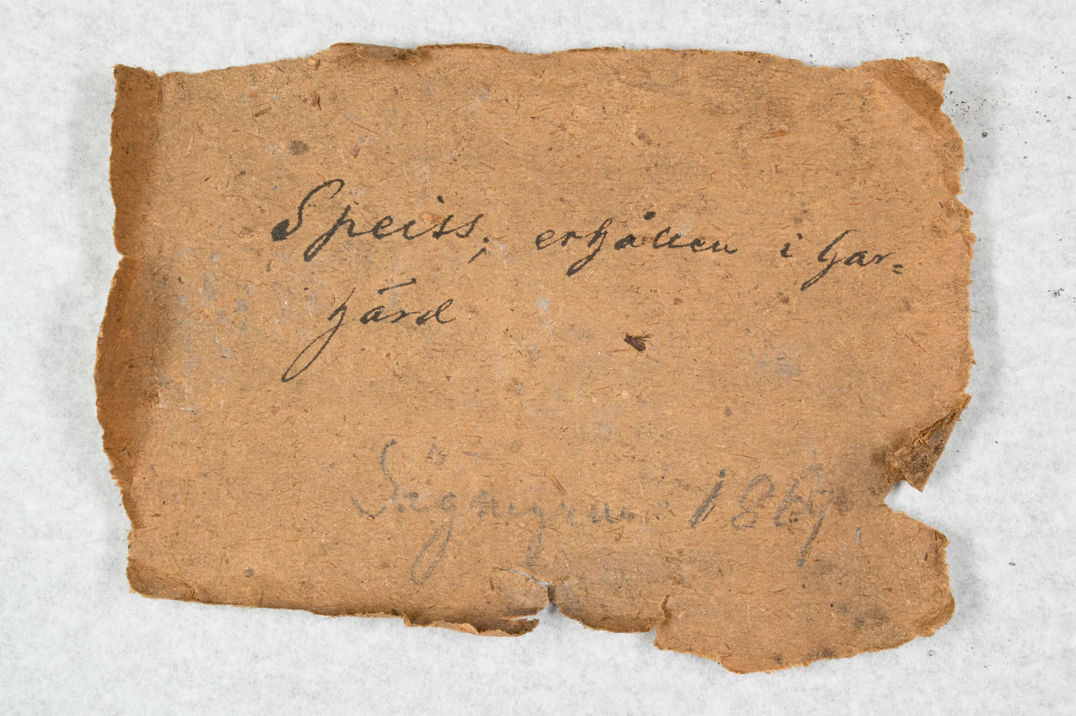 Fem materialprover, fyra svarta och ett vit och gråaktigt. Tillhörande lapp med notering: "Speiss, erhållen i Garhärd, Sågmyra 1869".