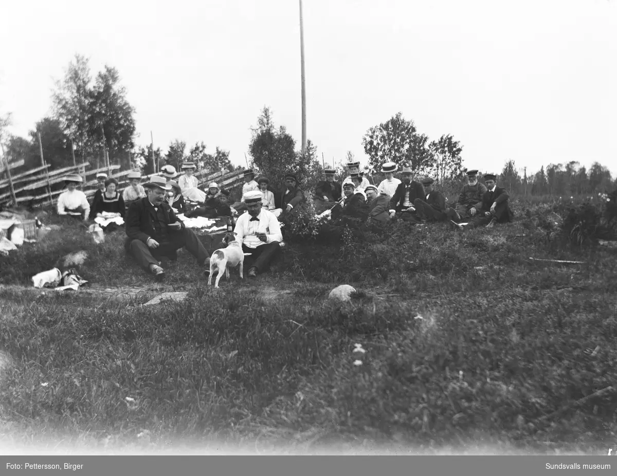 Gruppbild med en stor grupp kvinnor och män samt ett par barn som sitter i gräset framför en gärdesgård.