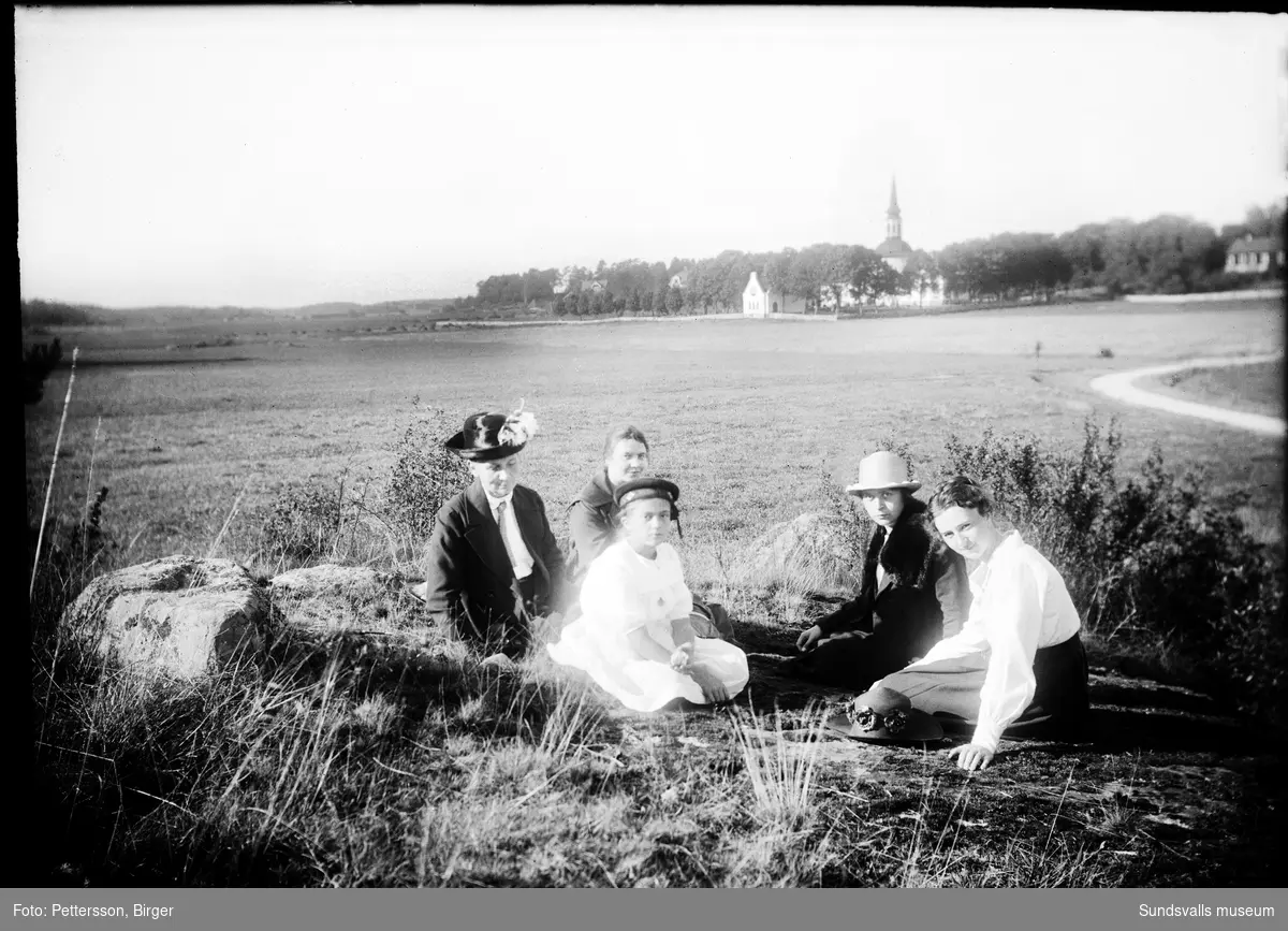 Fyra kvinnor och en flicka sitter i gräset på landsbygden. I bakgrunden syns en kyrka.