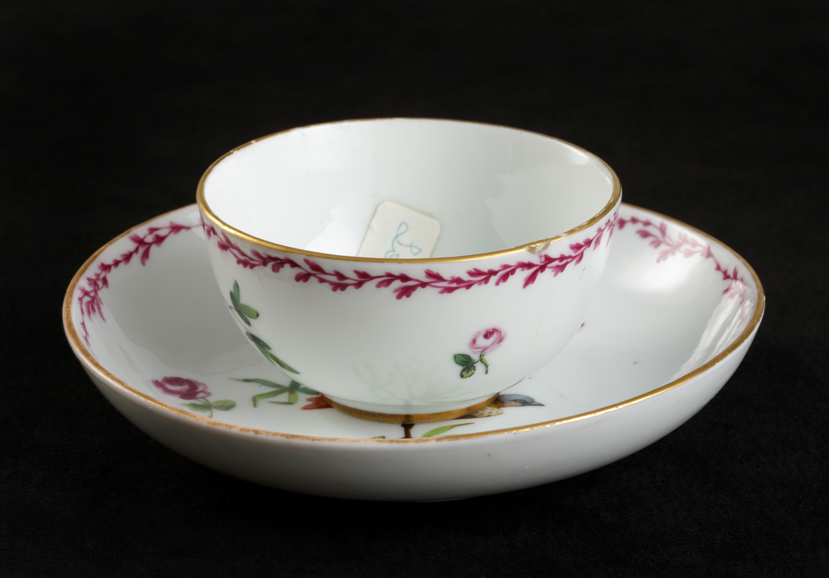 Kopp med fat, av porslin. Skålformad. 
Dekorerad med fågel på trädgren och strödda blommor. Längs kanterna lagergirland i rosa och guld.

Inskrivet i huvudbok 1967.