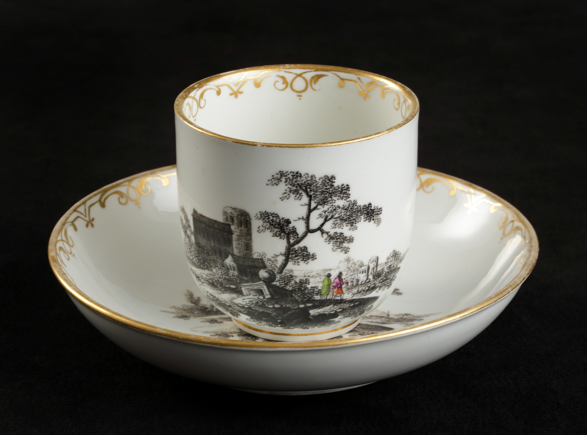 Kopp med fat av porslin. Koppen i bägarform. Fatet skålformigt. 
Dekorerad med ruinlandskap i svart camaïeu, med färglagda figurer. 
Kring kanterna spetsliknande bårder i guld. 

Inskrivet i huvudbok 1967.